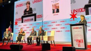 Woman speaks at a podium with four award winners sitting in chairs to her left.